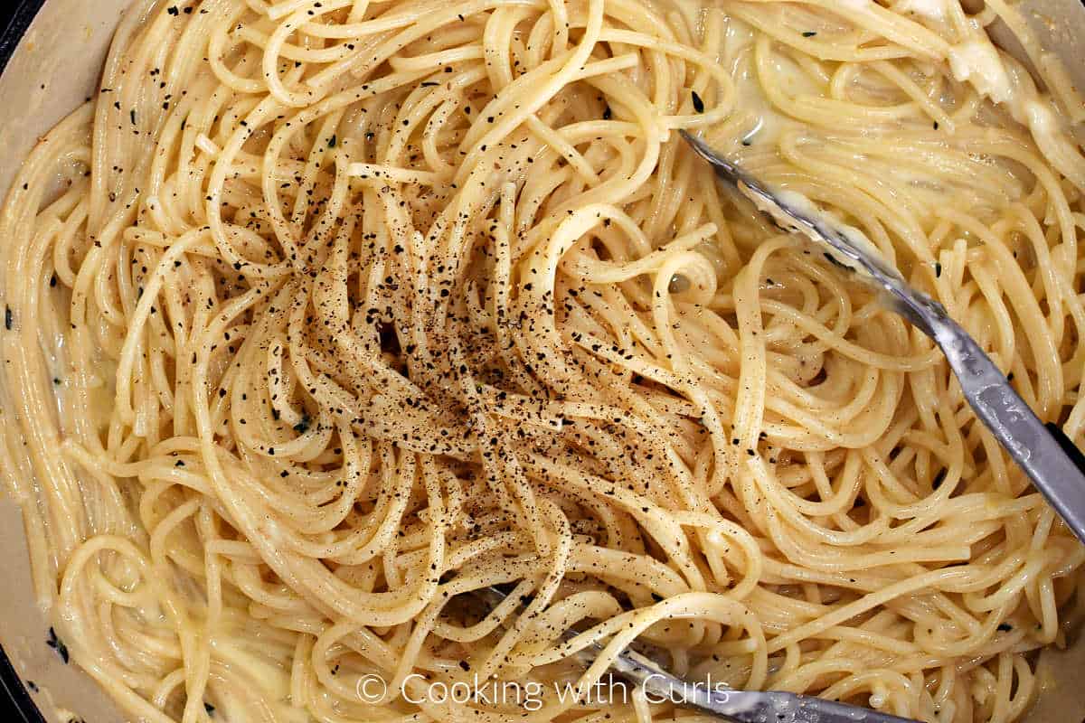 Cooked spaghetti in skillet with black pepper and tongs.