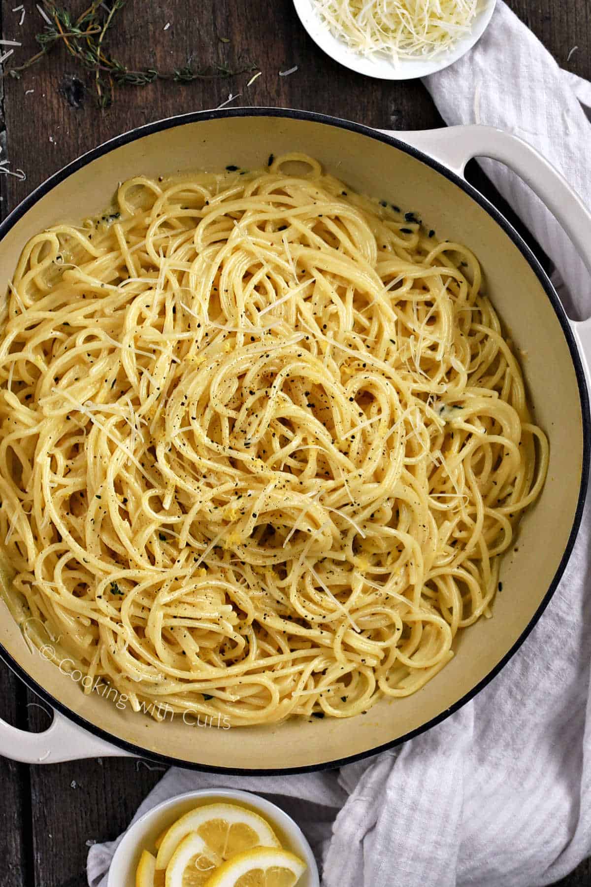 Looking down on spaghetti al limone in a skillet with lemon slices and parmesan cheese in bowls on the side.