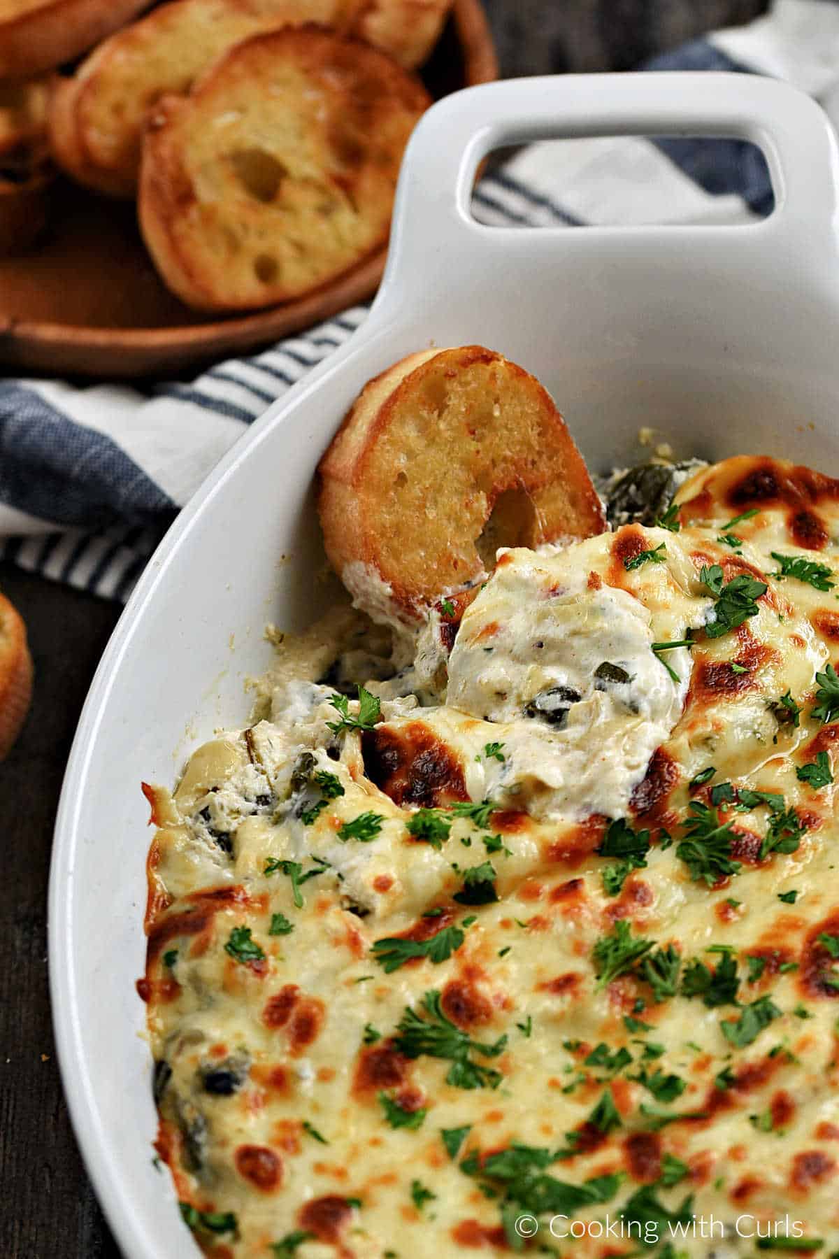 An oval baking dish filled with creamy spinach artichoke dip scooped with a bread crostini in the upper left corner. 