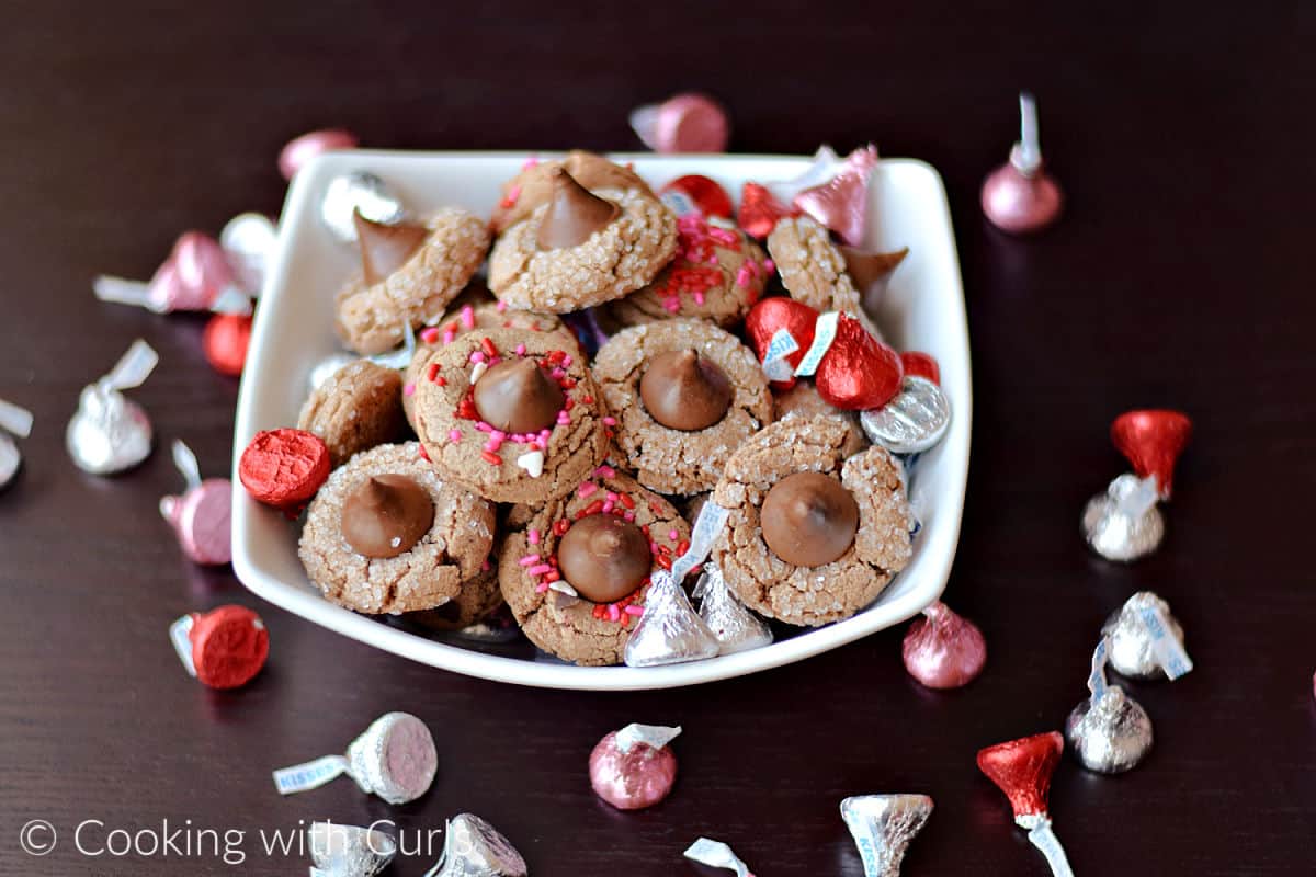 A bowl filled with of Chocolate Peanut Butter Blossom Cookies and pink, red and silver wrapped Hersheys Kisses.