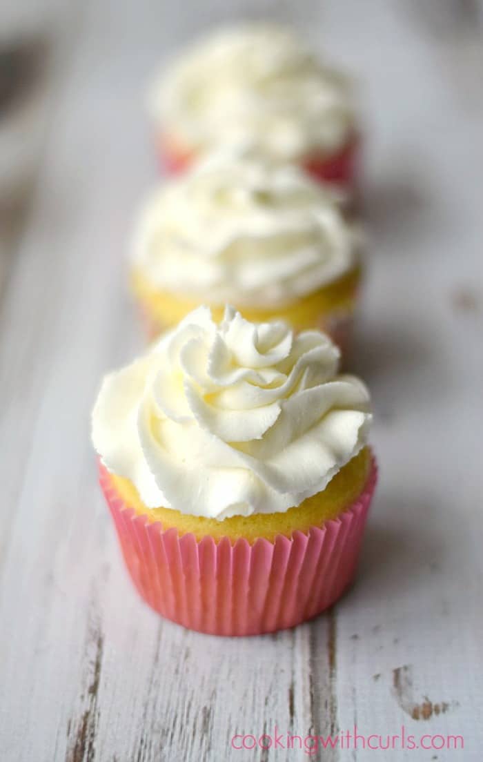Three white frosting covered cupcakes with pink and yellow wrappers in a row on a white background.