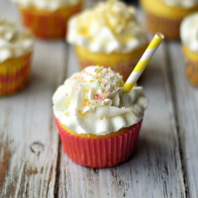 A white frosting topped cupcake with a pink wrapper and yellow striped straw sitting in front of six additional cupcakes on a white background.