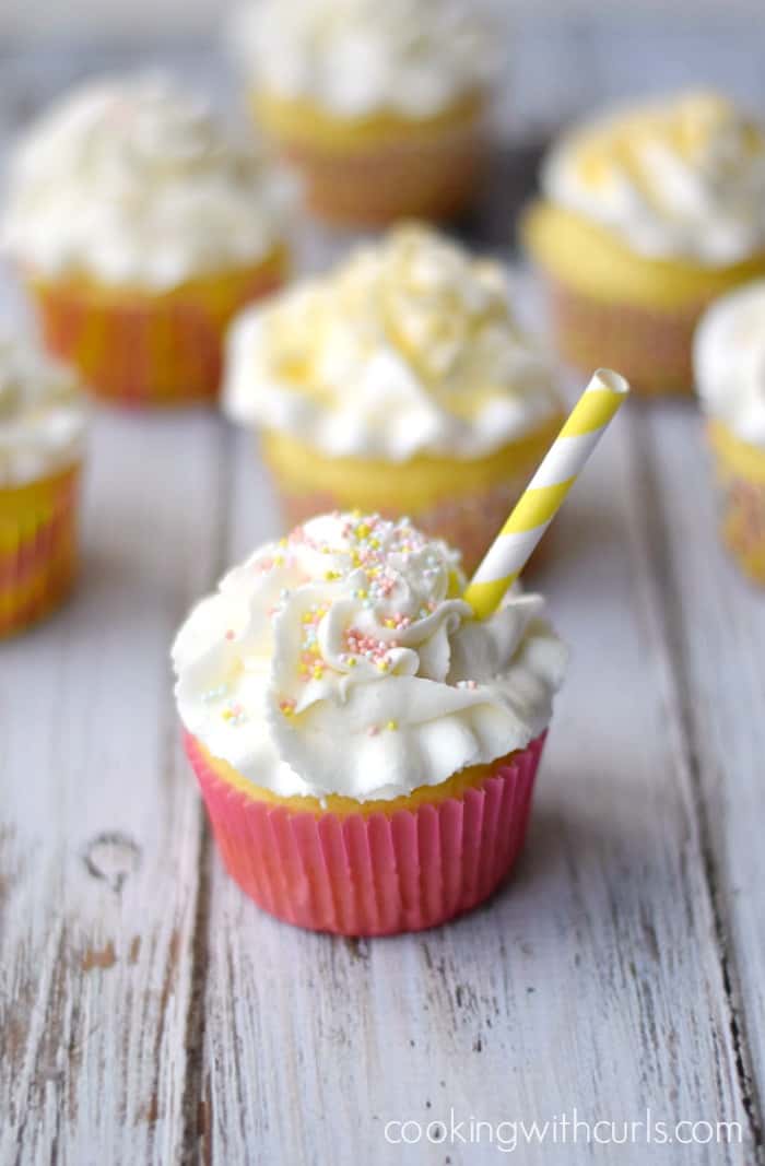 a white frosting topped cupcake with a pink wrapper and yellow striped straw sitting in front of six additional cupcakes on a white background