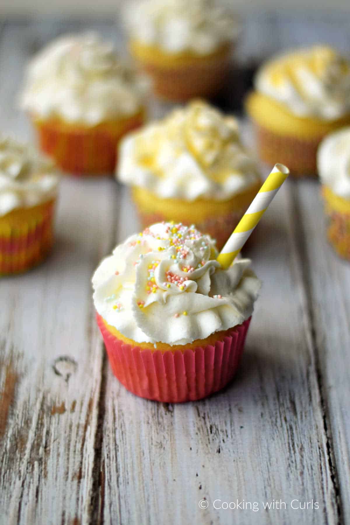 A white frosting topped cupcake with a pink wrapper and yellow striped straw sitting in front of six additional cupcakes on a white background.