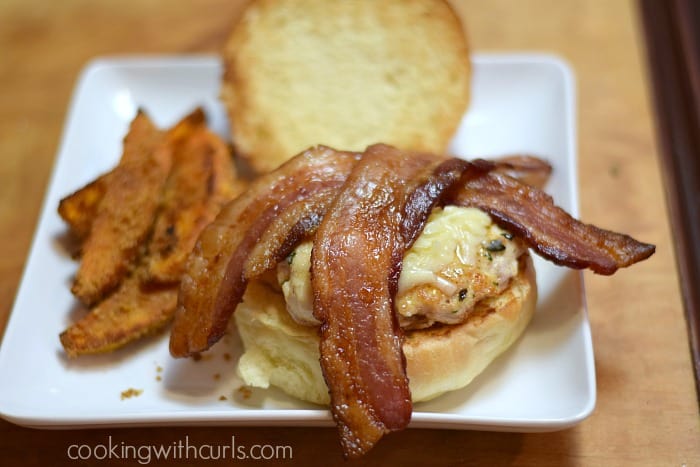 A melted cheese topped chicken breast topped with two slices of bacon on the bottom half of a bun with sweet potato fries on a white plate.