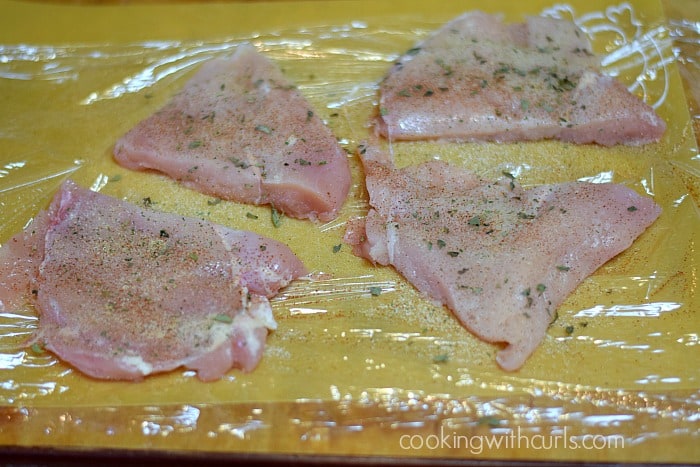 Four chicken breasts on plastic wrap covered in seasonings.