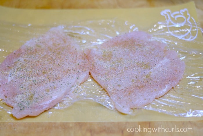 Two seasoned chicken breasts on a sheet of plastic wrap.