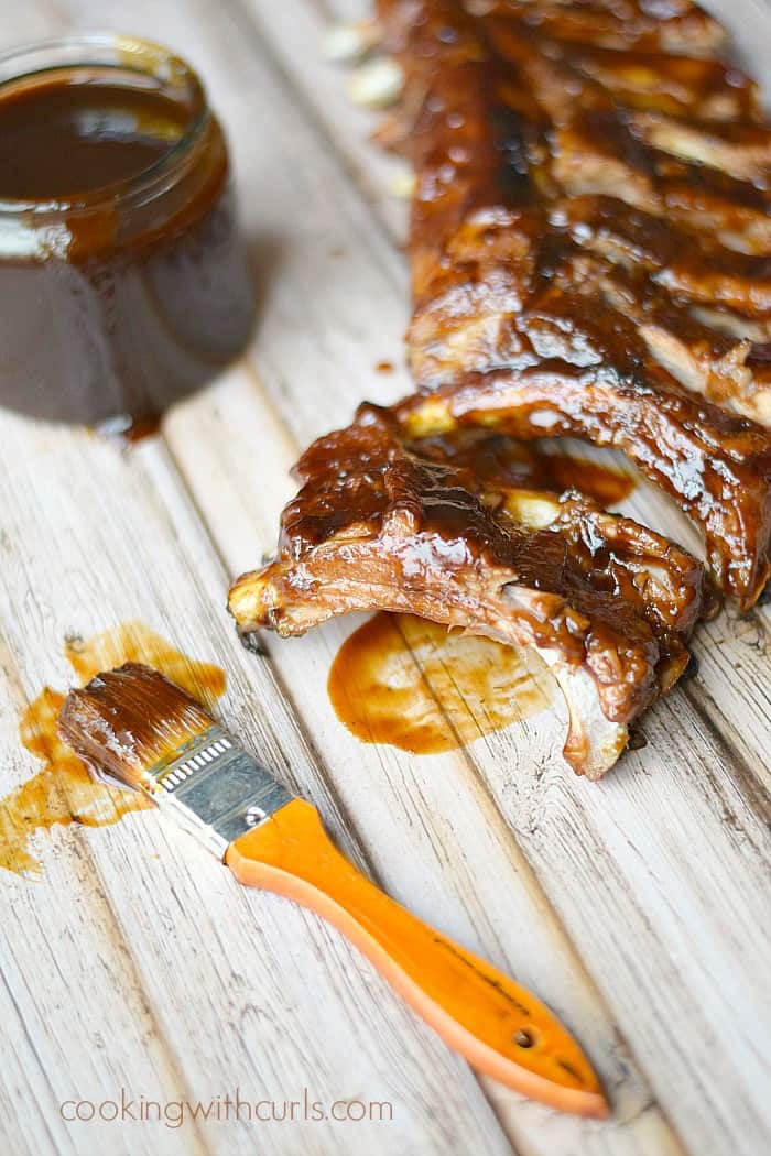 a rack of barbecue ribs with a sauce covered brush and a glass jar of sauce on a white wooden background