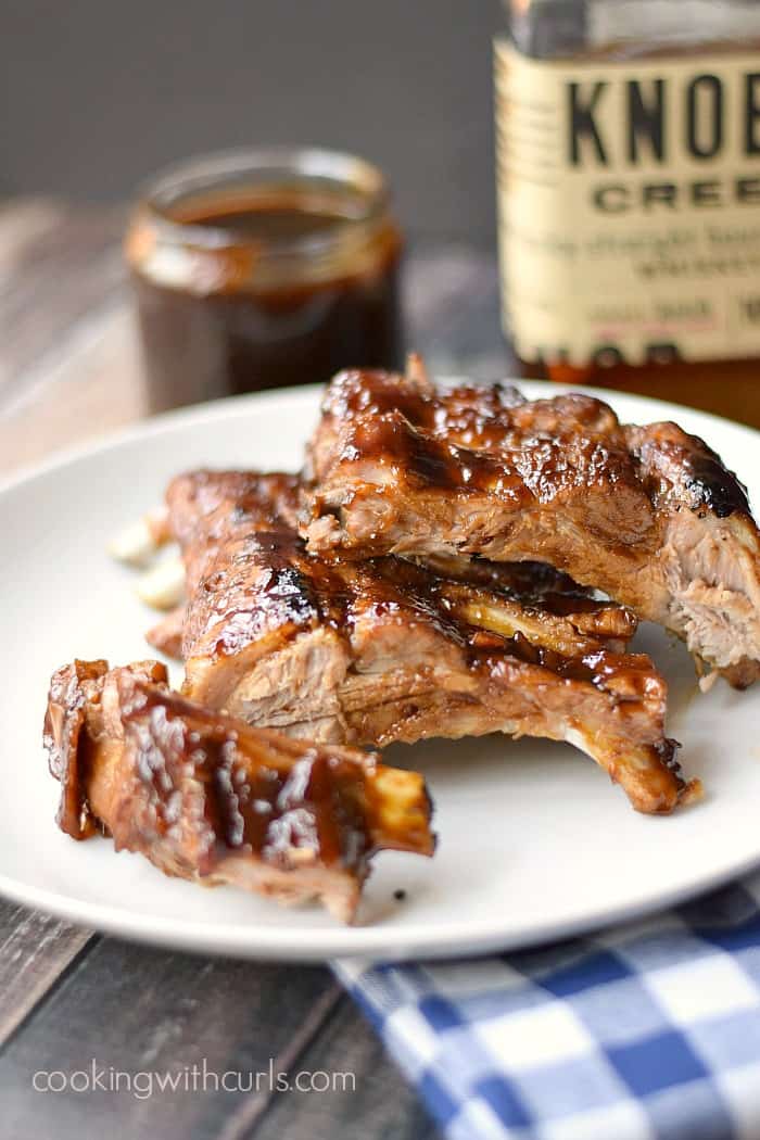 Barbecue ribs on a white plate sitting on a blue checkered napkin with a jar of barbecue sauce and bottle of bourbon in the background.