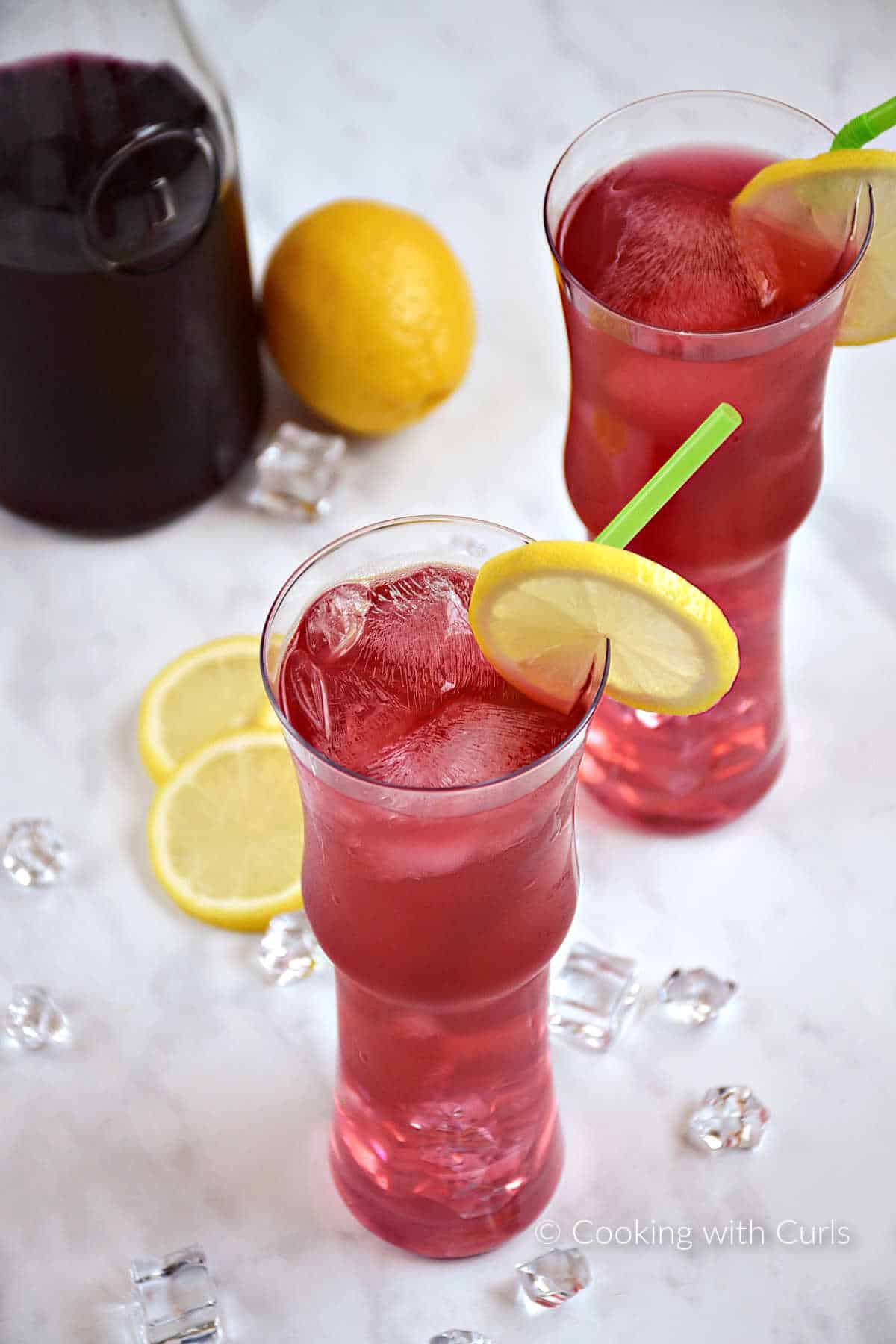 Two tall glasses filled with ice cubes and passion tea lemonade with a lemon slice garnish with a jar of passion tea in the background.