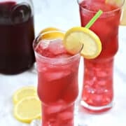 Two tall glasses filled with ice cubes and passion tea lemonade with a lemon slice garnish.