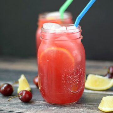 two glass mason jars filled with cherry lemonade with one blue and one green straw with lemon wedges and fresh cherries surrounding them on a wooden board
