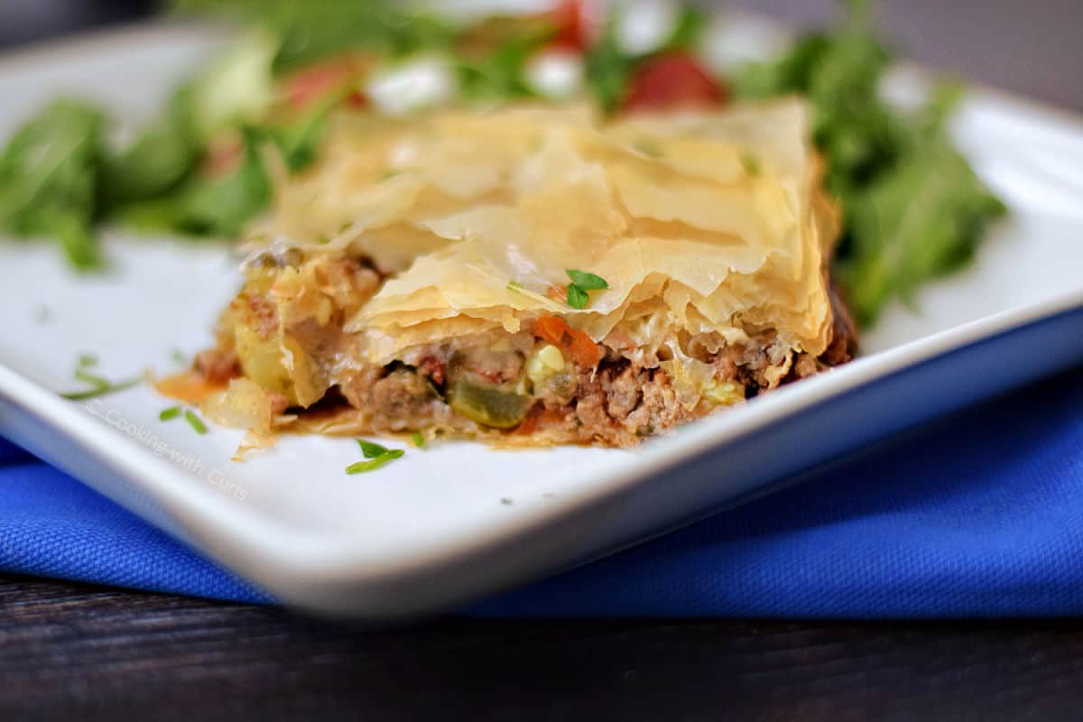 A slice of Greek phyllo meat pie on a plate with lettuce salad.