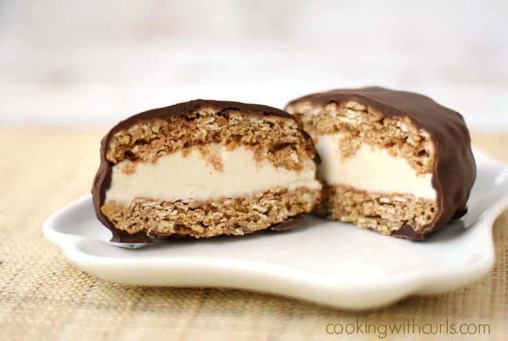 Vanilla ice cream surrounded by two oatmeal cookies and coated in rich chocolate, cut in half and sitting on a small plate.