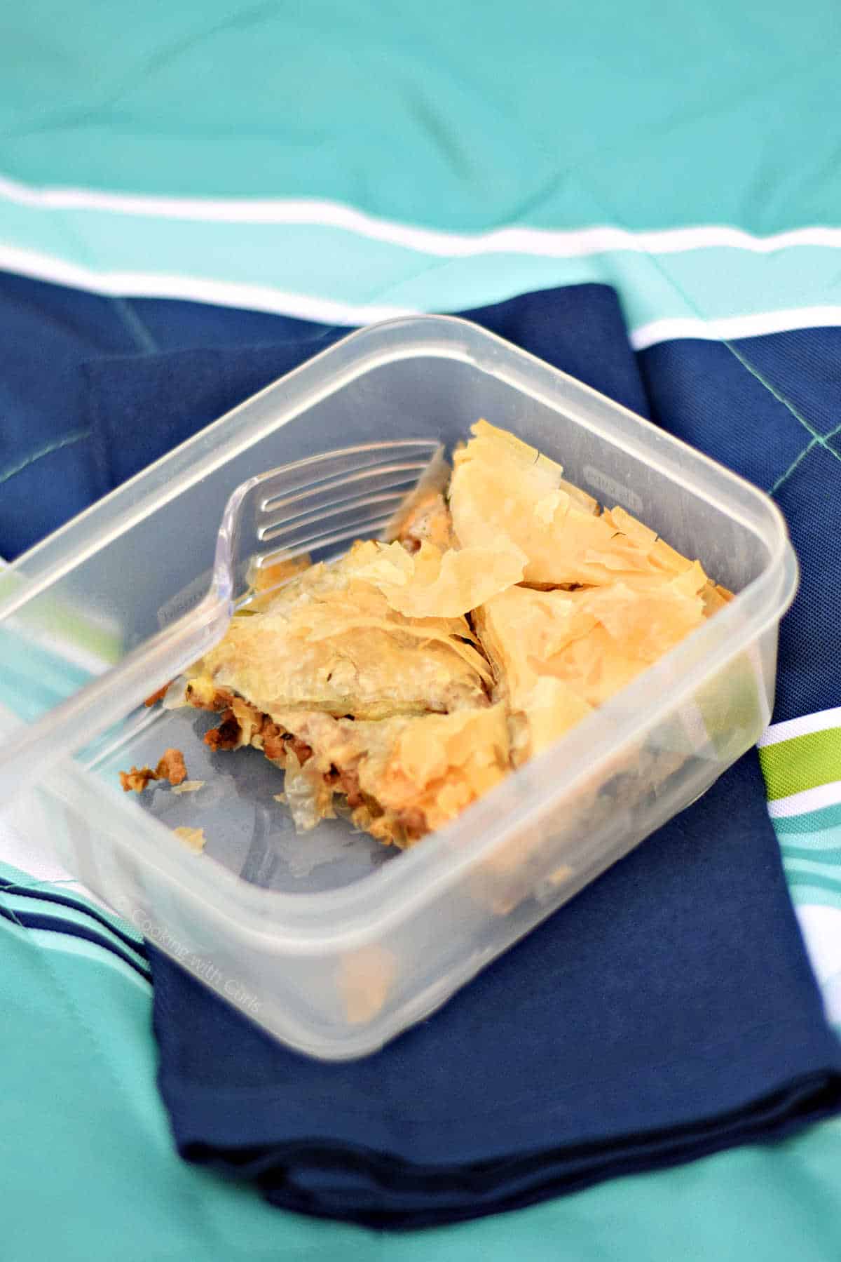 A piece of Greek phyllo meat pie in a plastic picnic container on a blue blanket.