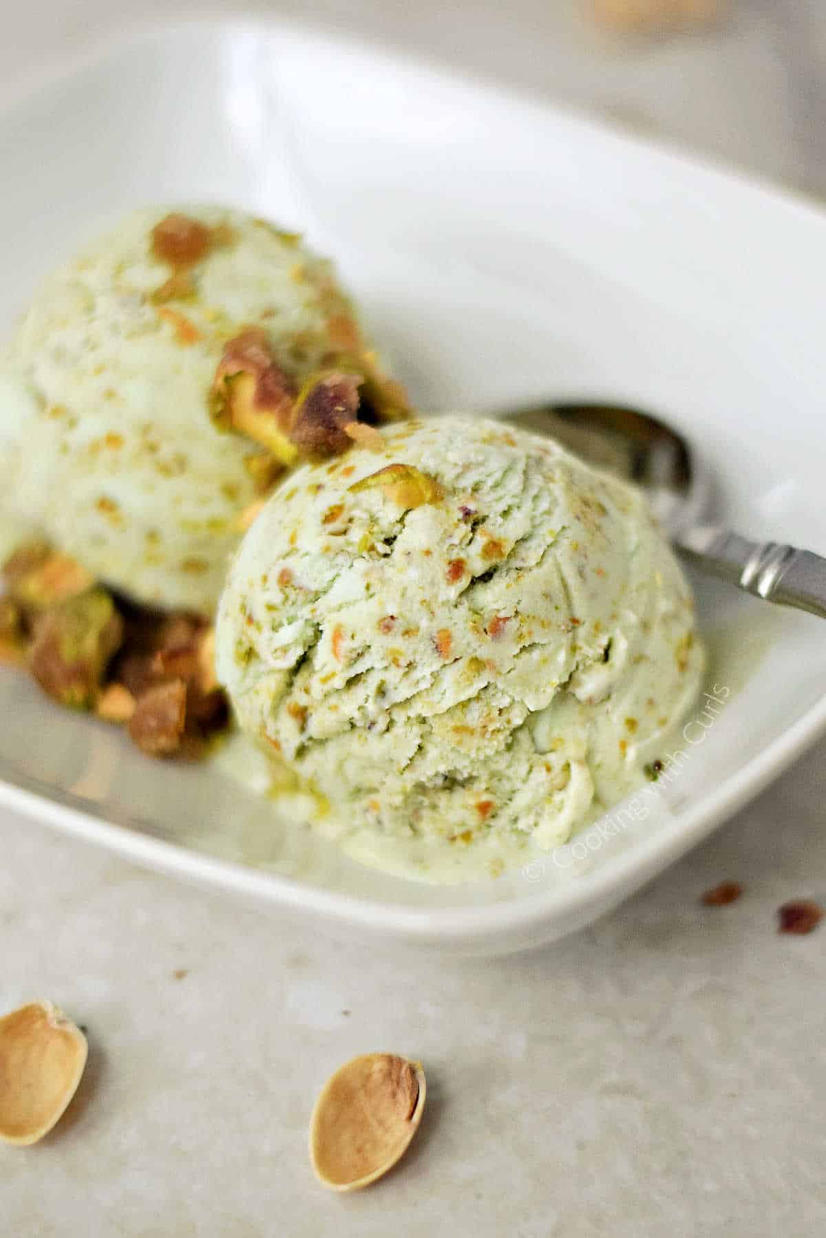 Pistachio Ice Cream topped with Pistachio Praline in a white rectangle dish with a spoon on the side.