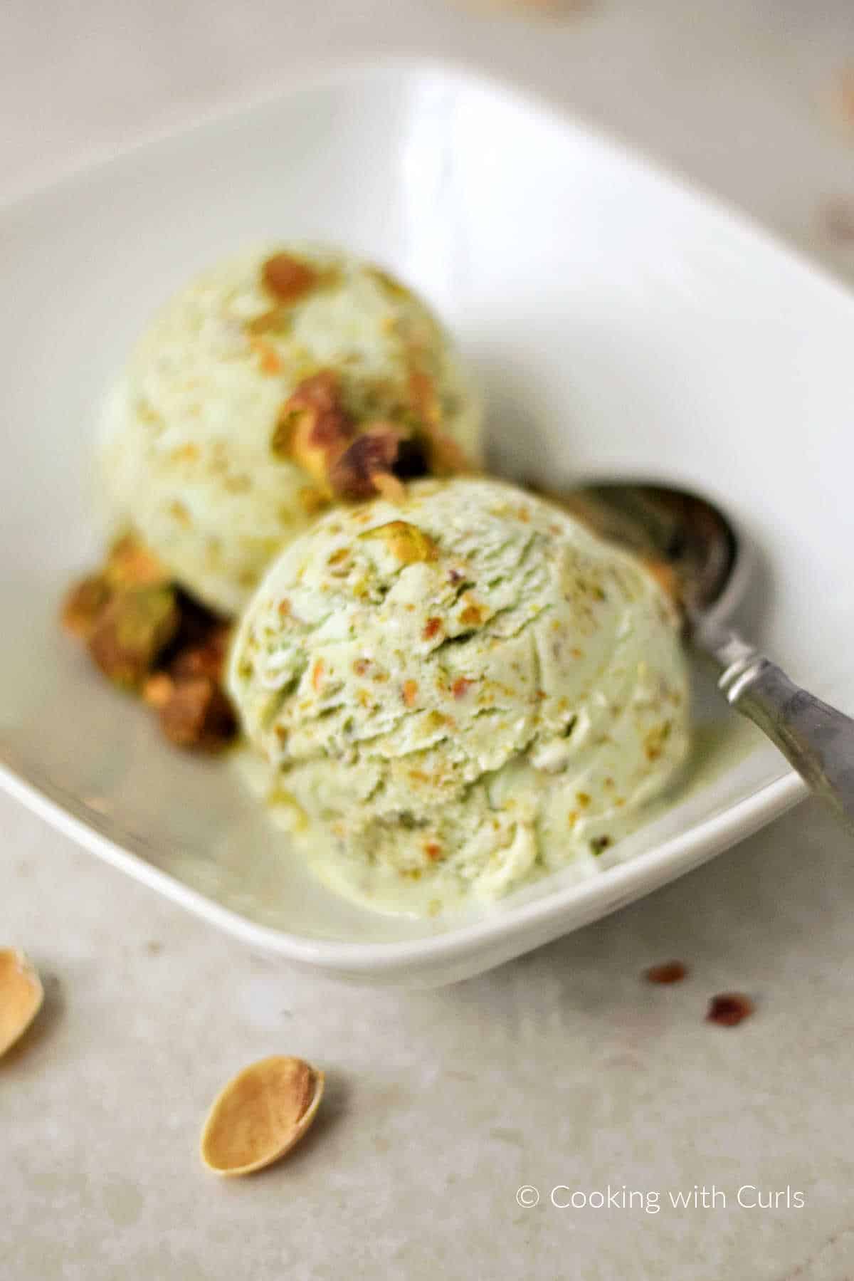 Two scoops of Pistachio Ice Cream with pistachio praline in a white bowl with a spoon on the side.