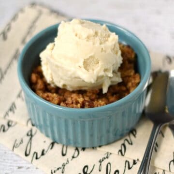 Rhubarb Crisp topped with vanilla ice cream in a small blue bowl sitting on a French script napkin with a spoon on the side.