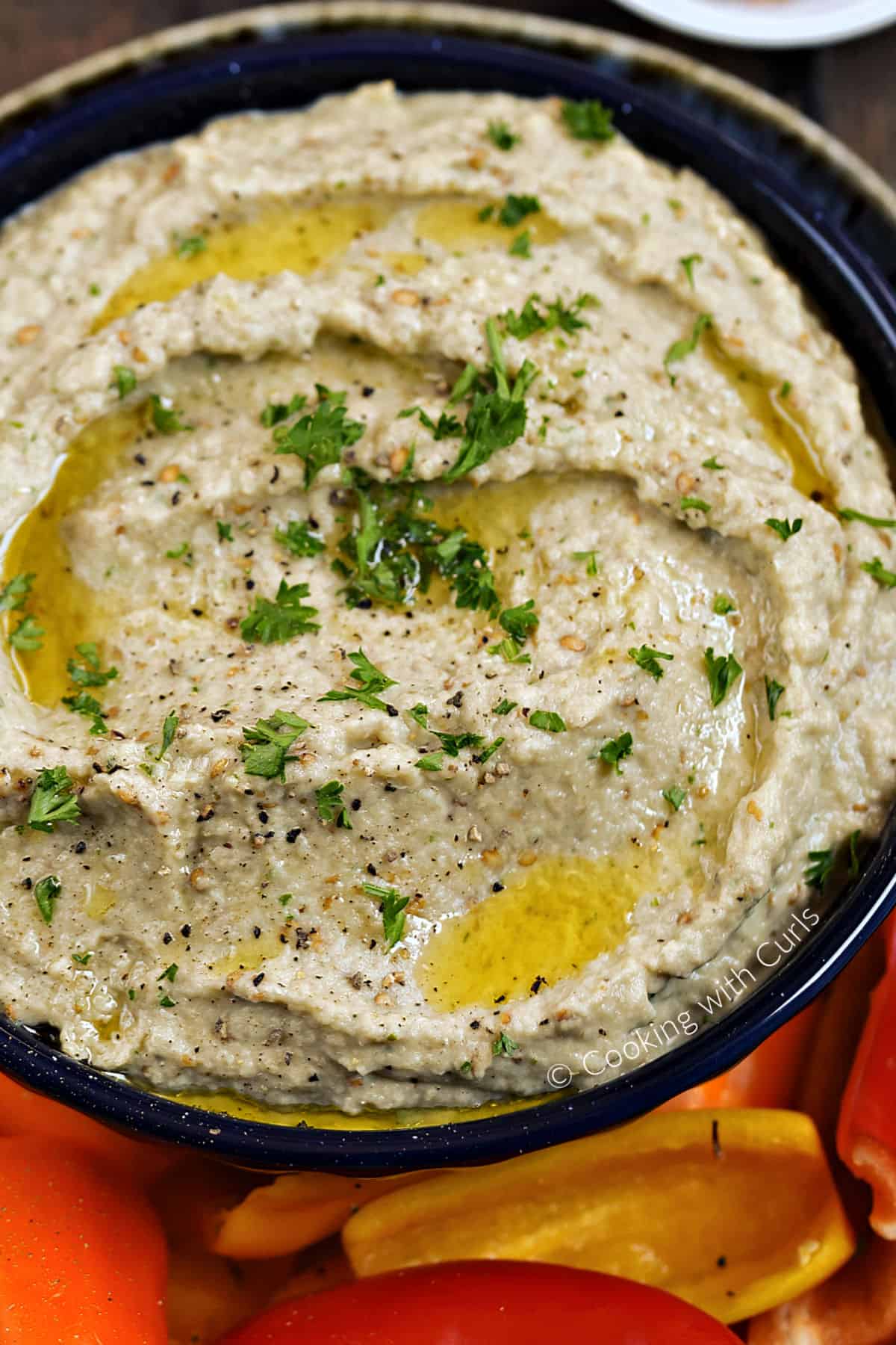 Looking down on a bowl of roasted Greek Eggplant Dip drizzled with olive oil and sprinkled with chopped parsley on a platter filled with sliced bell pepper.