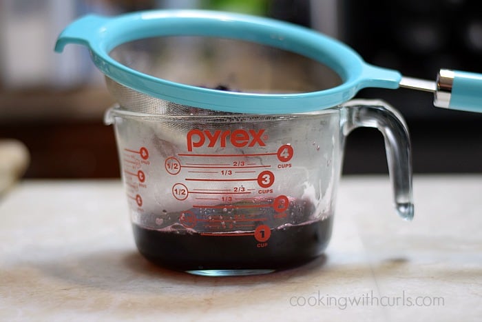 A blue strainer sitting on a large glass measuring cup filled with blackberry syrup.