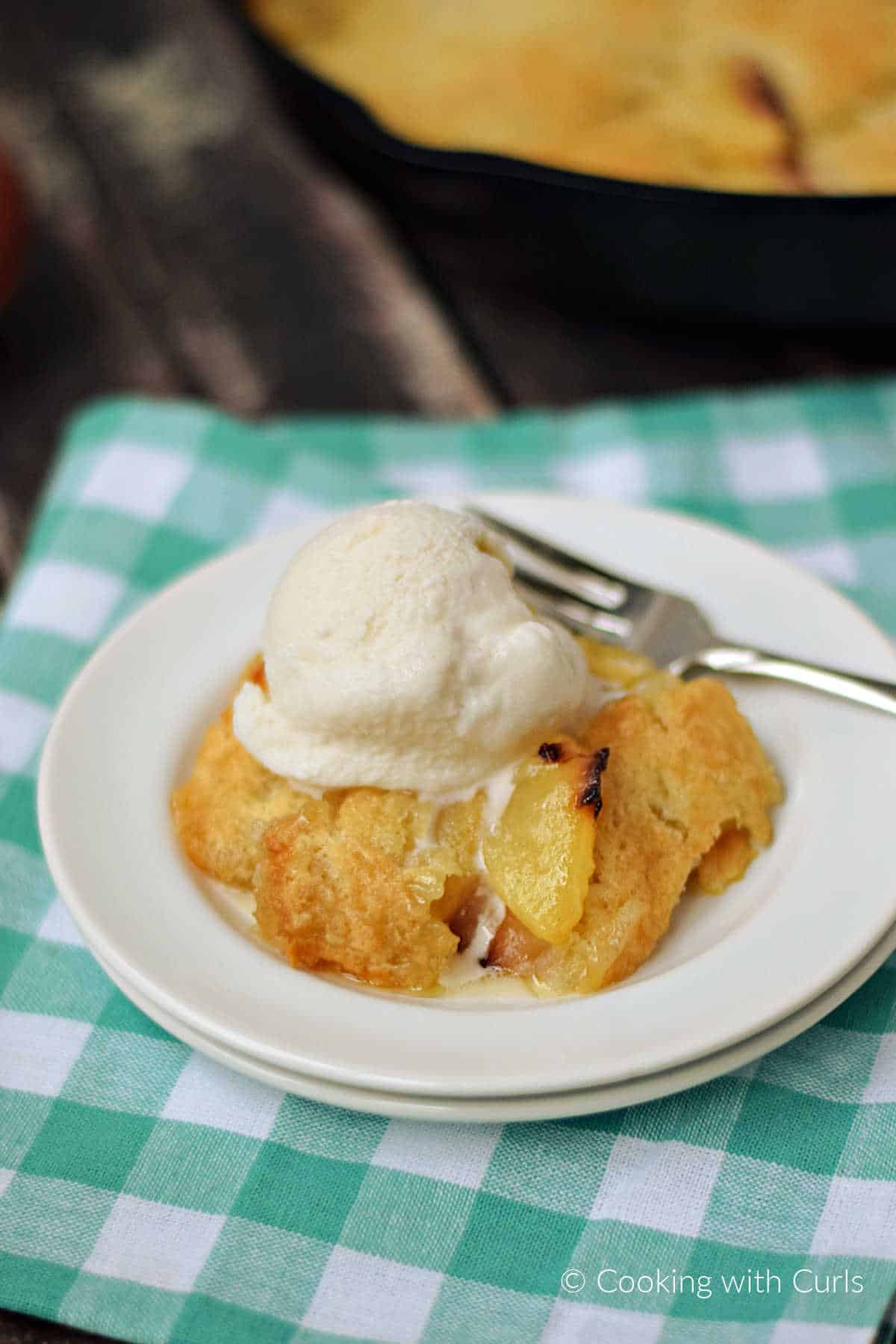 Peach cobbler on a plate topped with vanilla ice cream with a skillet of cobbler in the background.