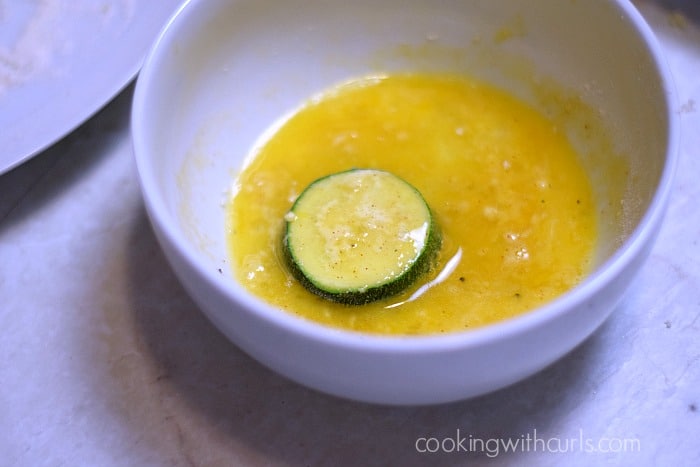 Flour coated zucchini slice in a bowl with beaten egg.