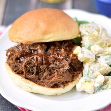 barbecue beef sandwich with a side of potato salad on a white plate that is sitting on a red and white check napkin