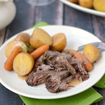 sliced pot roast, vegetables and gravy on a white plate sitting on a green napkin with another plate of pot roast in the background