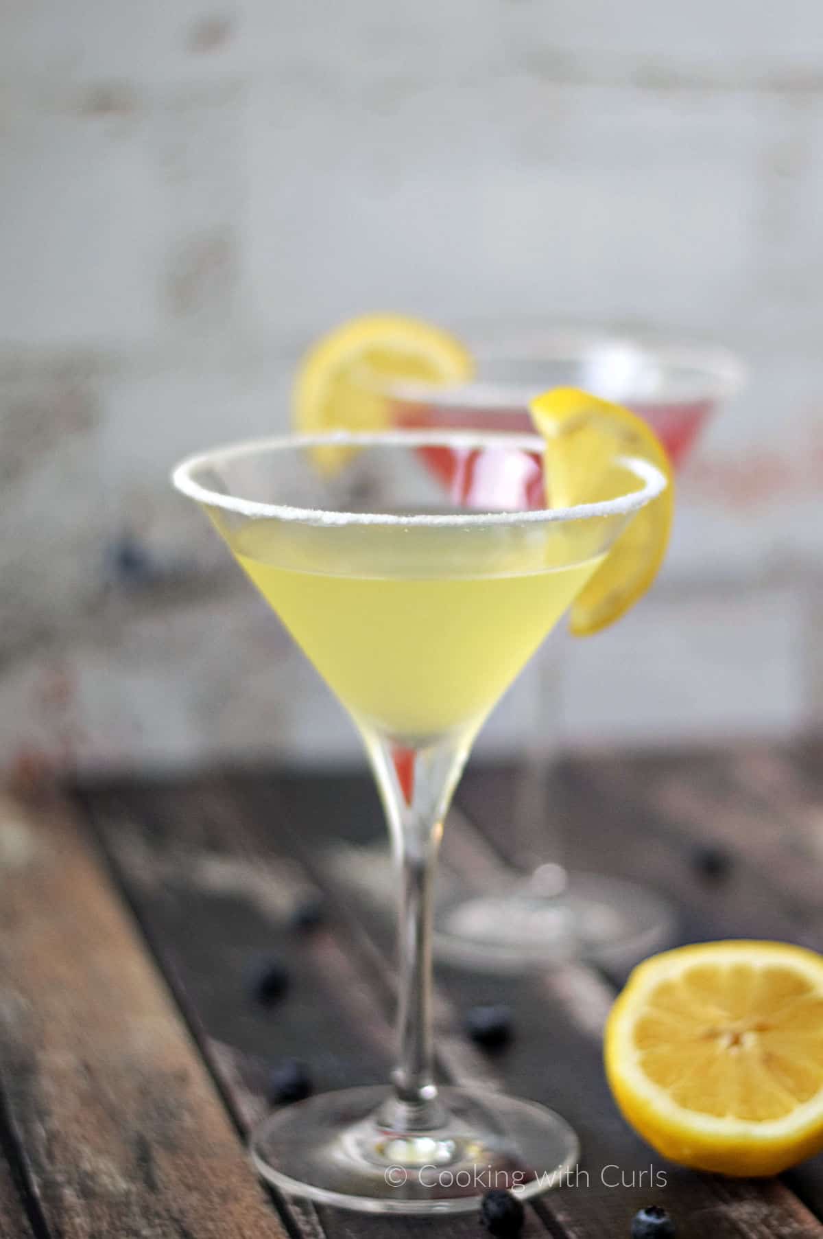 a lemon drop martini sitting in front of a huckleberry martini each garnished with a lemon slice.
