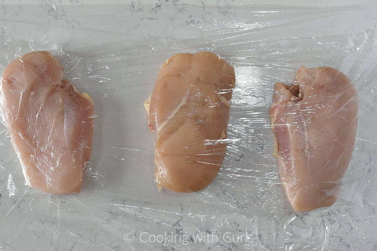 Three chicken breasts wrapped in plastic wrap on the counter.