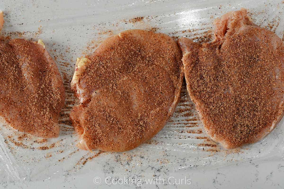 Three chicken breasts coated with southwest seasoning on a sheet of plastic wrap.