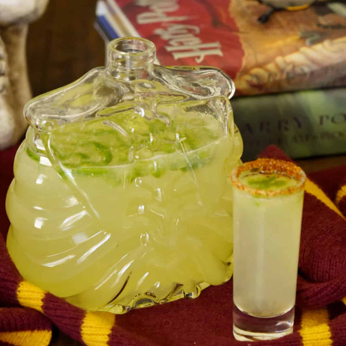 A skeleton shaped decanter filled with spicy jalapeno margarita recipe in front of a stack of Harry Potter books with a filled shot glass in front. 