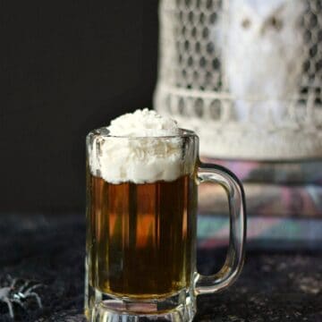 butter beer cocktail in a glass mug in front of a stack of harry potter books topped with a white owl in a white cage