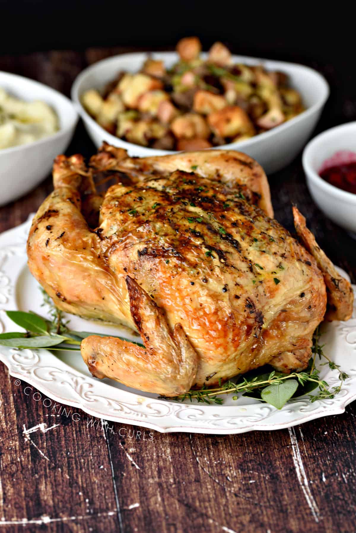 A whole roast chicken on a white platter with bay leaves and thyme with bowls of stuffing and mashed potatoes in the background.