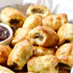 Puff pastry sausage rolls piled up in a paper lined basket with dipping sauce on the left side.