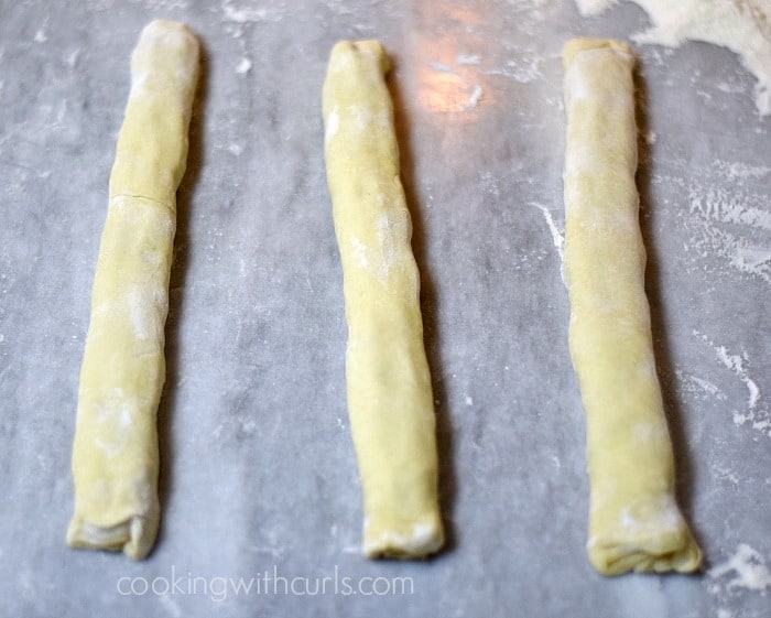 Three strips of Puff Pastry rolled around Sausage filling on a marble board.