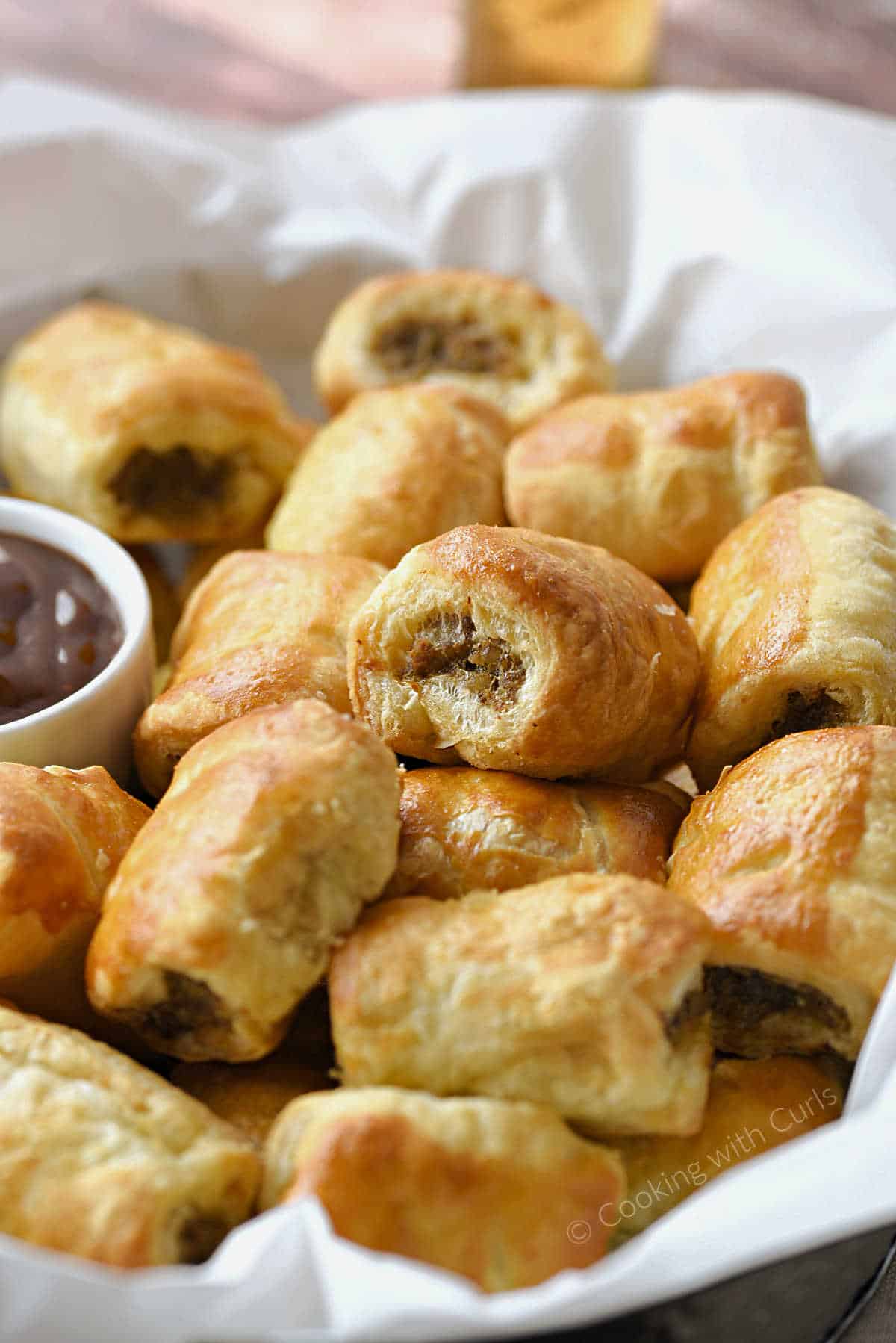 Puff pastry sausage rolls piled up in a serving basket with a side of dipping sauce.