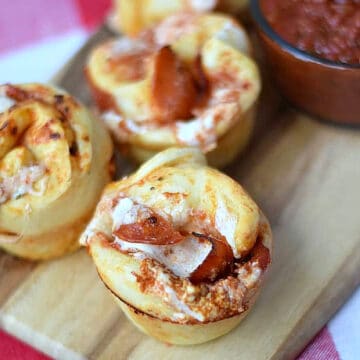 Three Pepperoni Pizza Muffins on a serving board with a bowl of pizza sauce in the background. 