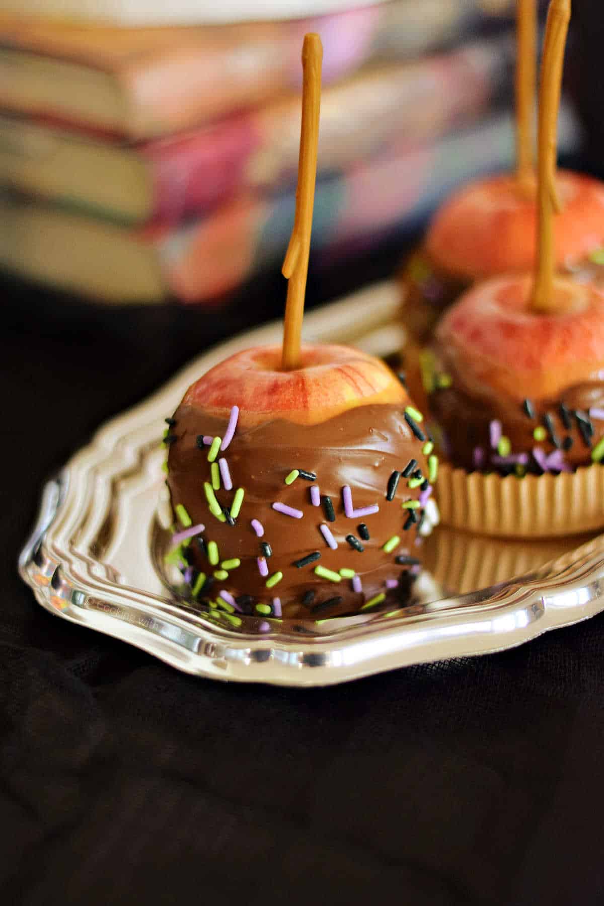 Three chocolate covered caramel apples with halloween sprinkles on a silver tray.