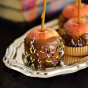 Three chocolate covered caramel apples with halloween sprinkles on a silver tray.