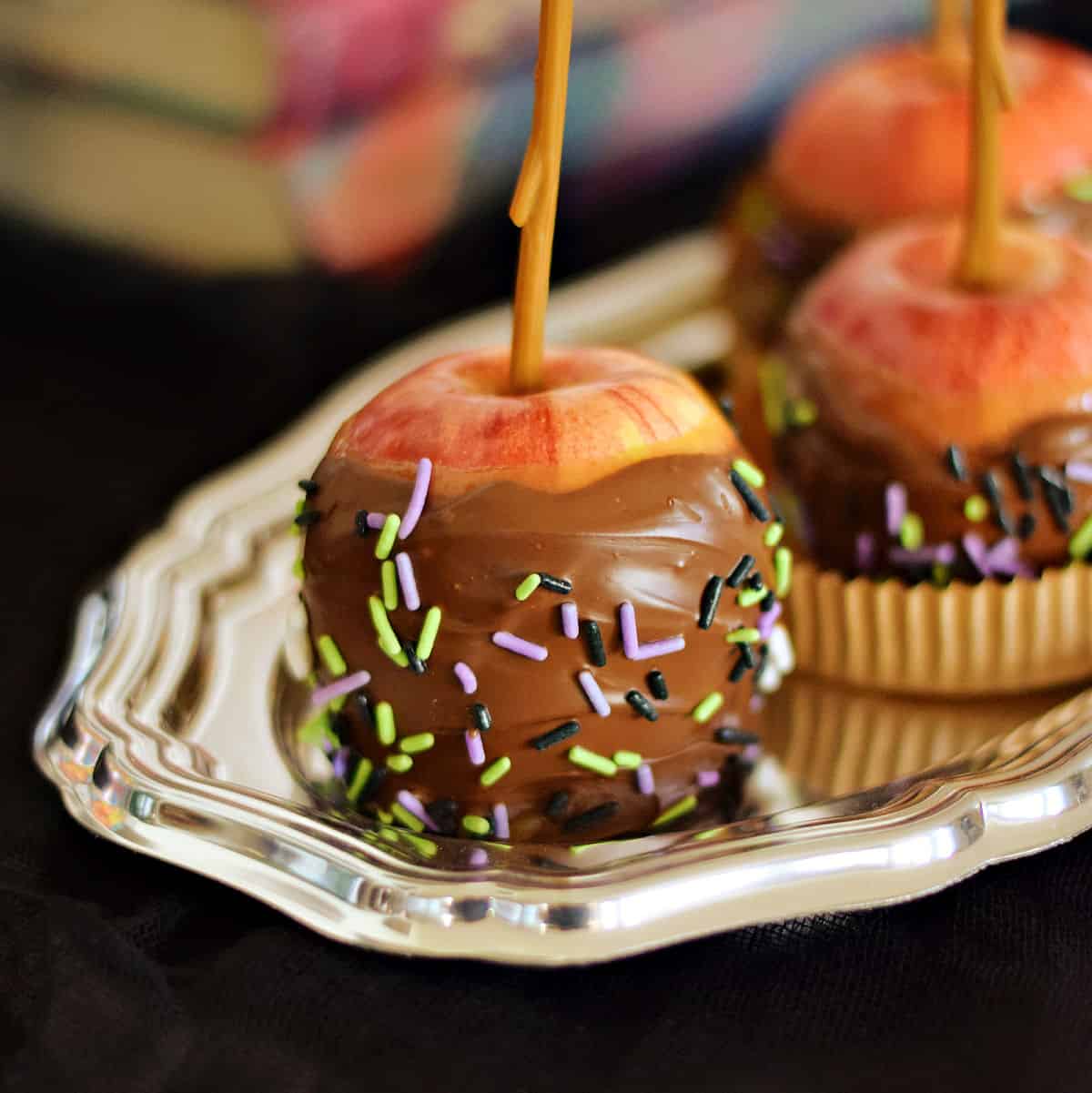 Three chocolate covered caramel apples with halloween sprinkles on a silver tray.