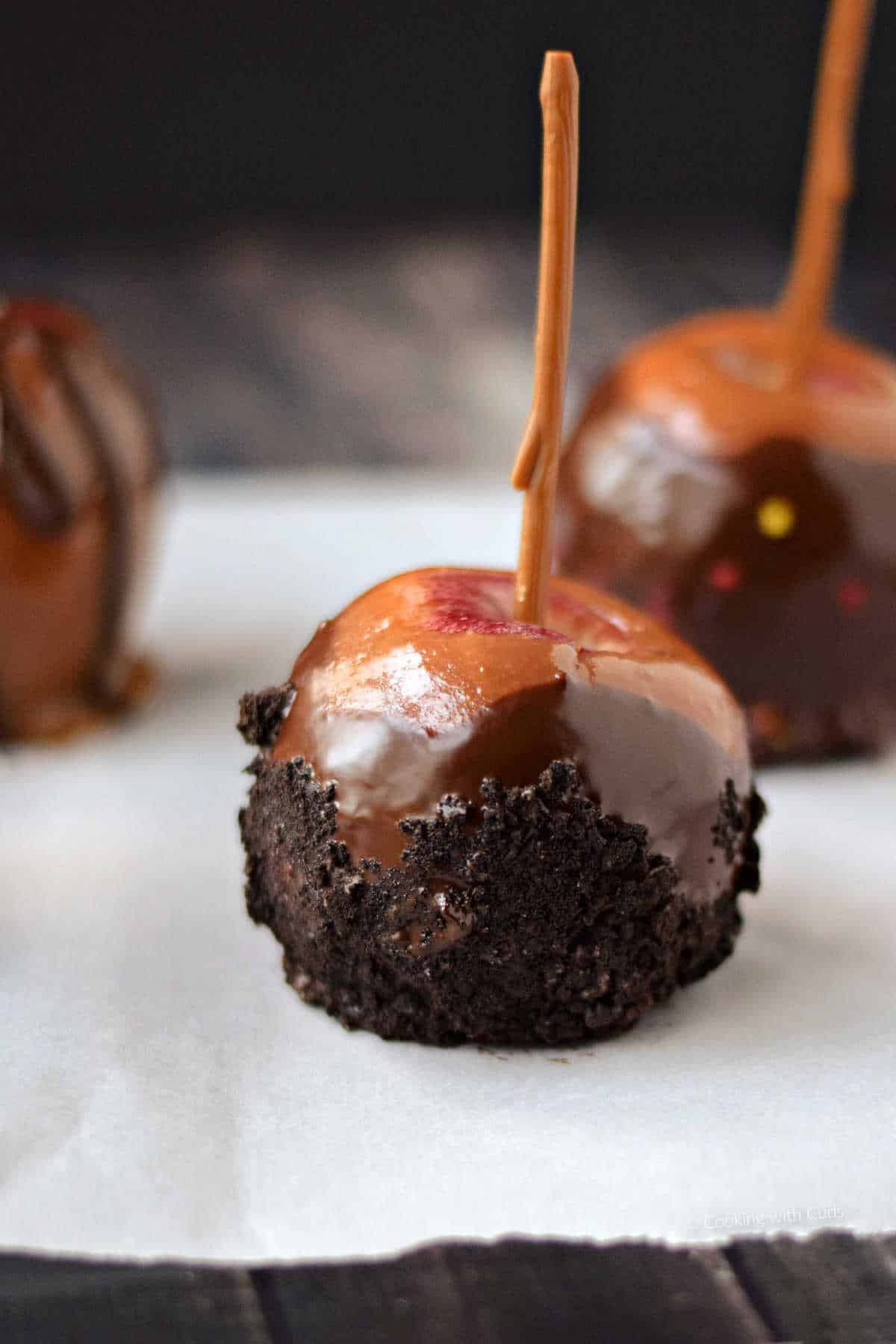 Three caramel apples with crushed oreos, sprinkles, and chocolate drizzle on parchment paper.