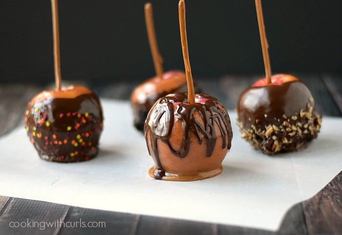 Four caramel apples on white parchment paper, each with different toppings
