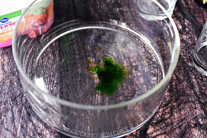green food coloring mixed with vodka in a large glass punch bowl with a carton of rainbow sherbet in the upper left corner. 