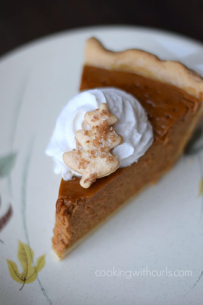 A slice of pumpkin pie topped with whipped cream and a sugar coated leaf pie crust cookie on a small plate.