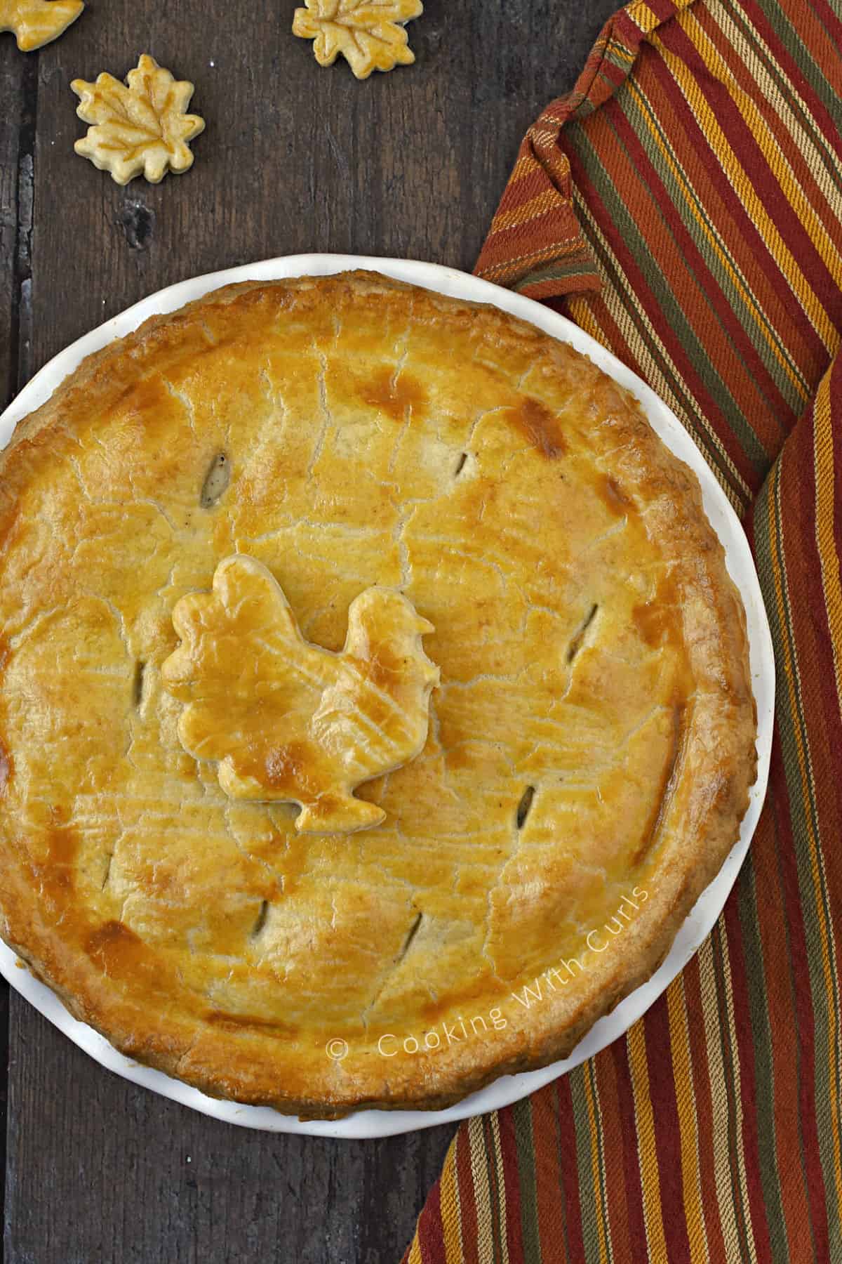 Looking down on a golden brown turkey pot pie with a turkey pie crust cut-out sitting on a fall colored striped towel. 