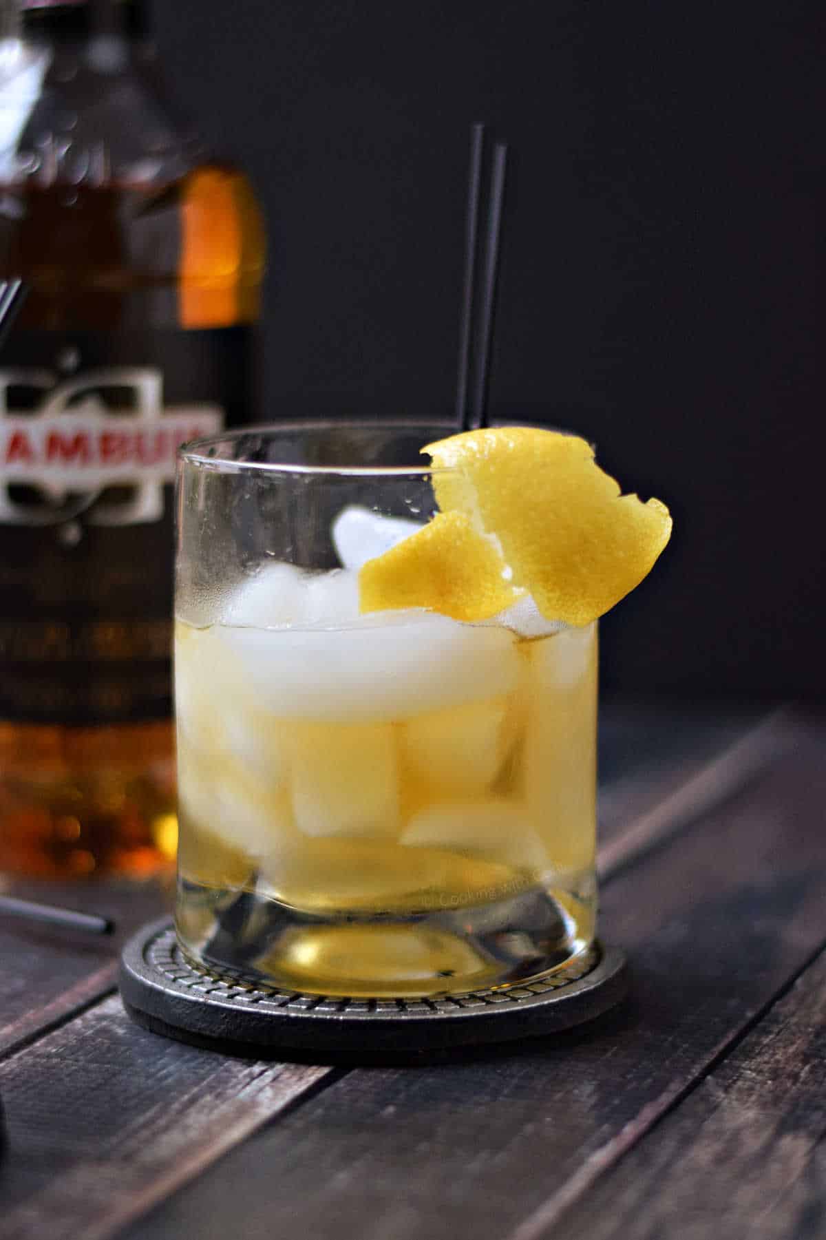 A rusty nail cocktail in a short glass filled with ice cubes and a lemon twist.