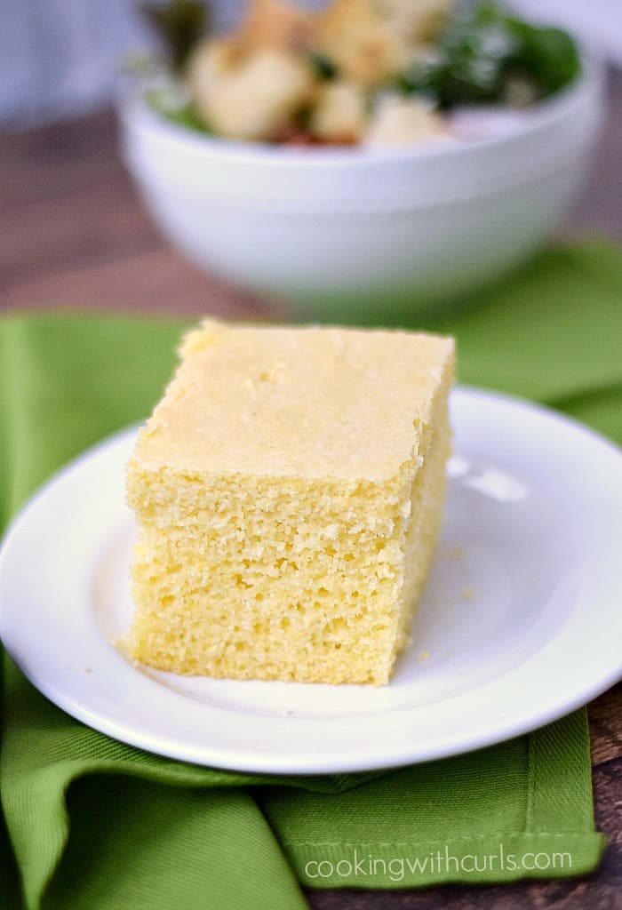 a slice of cornbread on a small, white plate sitting on a green napkin with a bowl of salad in the background