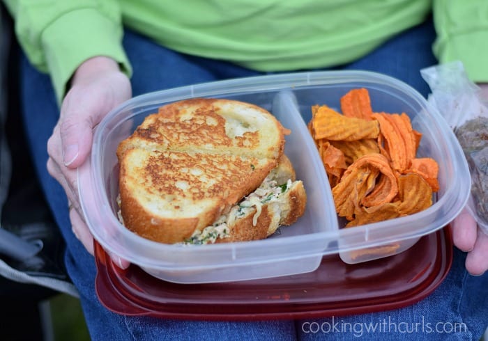 A woman's hands holding a plastic container with a gourmet tuna melt in one section and sweet potato chips in the other.