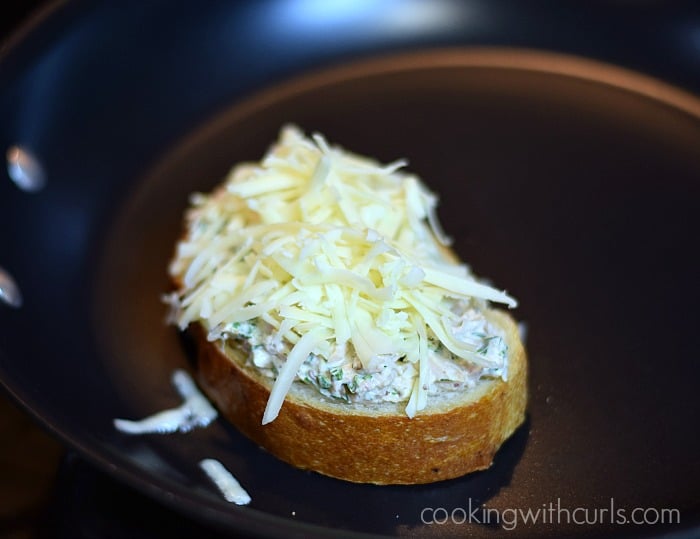 Tuna salad topped with grated cheese on a thick slice of sourdough bread in a non-stick skillet.